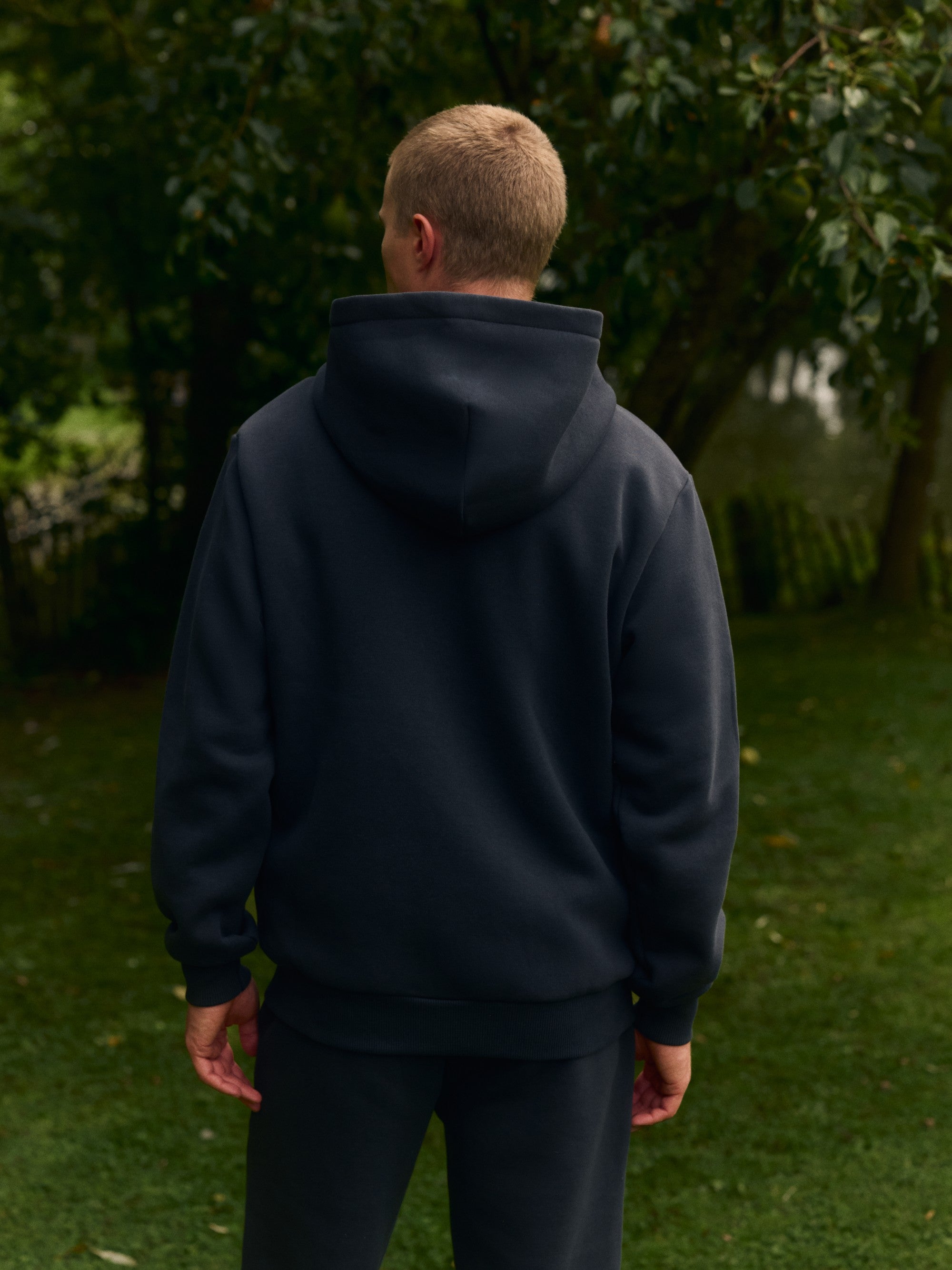 Back view of a man wearing a black CASPARA hoodie outdoors in a garden.